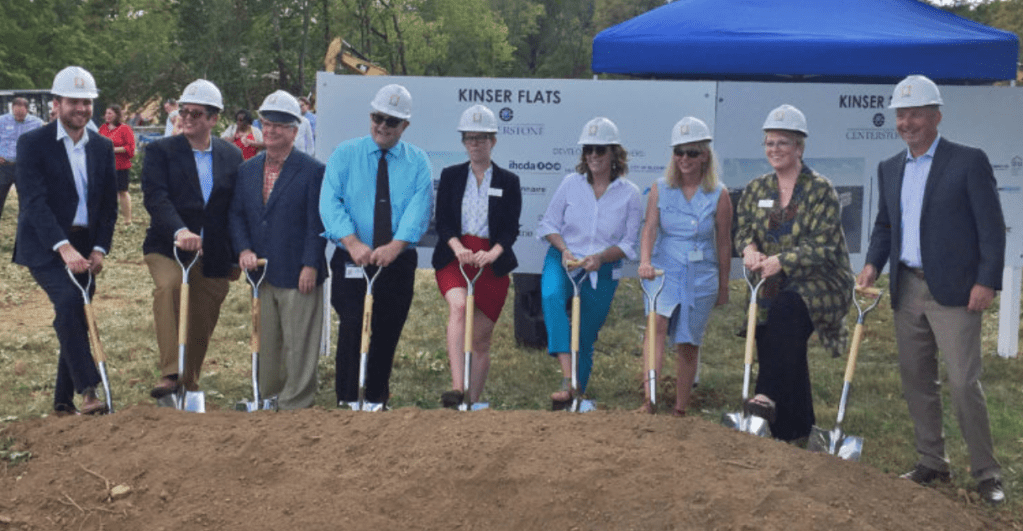 Nine people in white hard hats at the groundbreaking ceremony for Kinser Flats. Each person has a shovel placed into the dirt. Blue tent in background with sign saying "Kinser Flats."